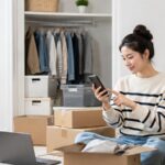 クローゼットと段ボール箱の前でスマホを使い、ミニクラとサマリーポケットを比較する女性